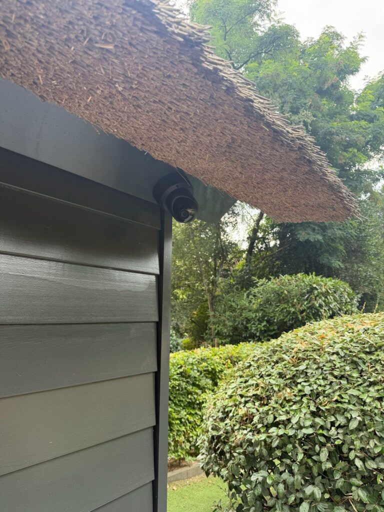 Camera onder de rietrand van een donkergroen tuinhuis, uitkijkend over de tuin