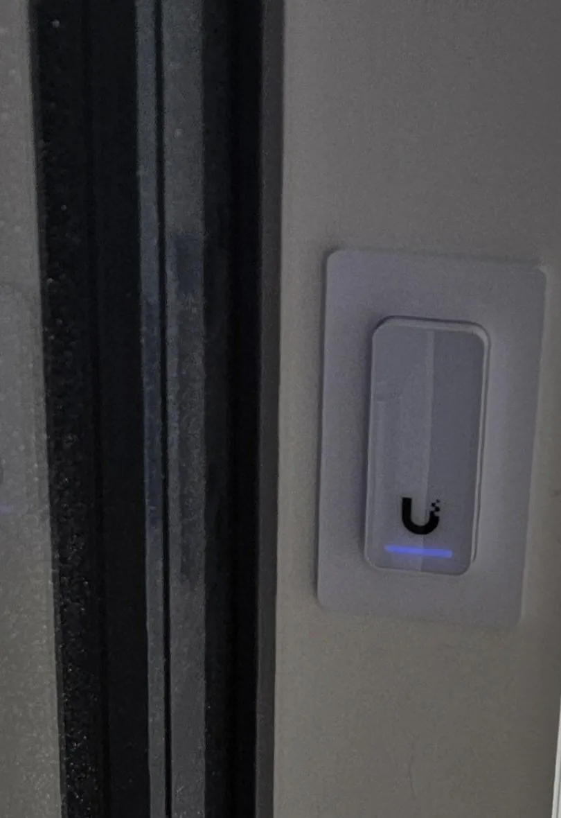 Toegangslezer met blauw lampje op een montageplaat naast een donker kozijn